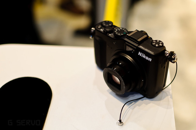 A black Nikon digital camera sits on a white surface with a black curved shape nearby.