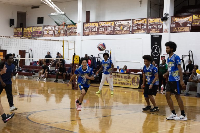 Five boys in blue jerseys play volleyball in a high school gym with spectators sitting on bleachers.