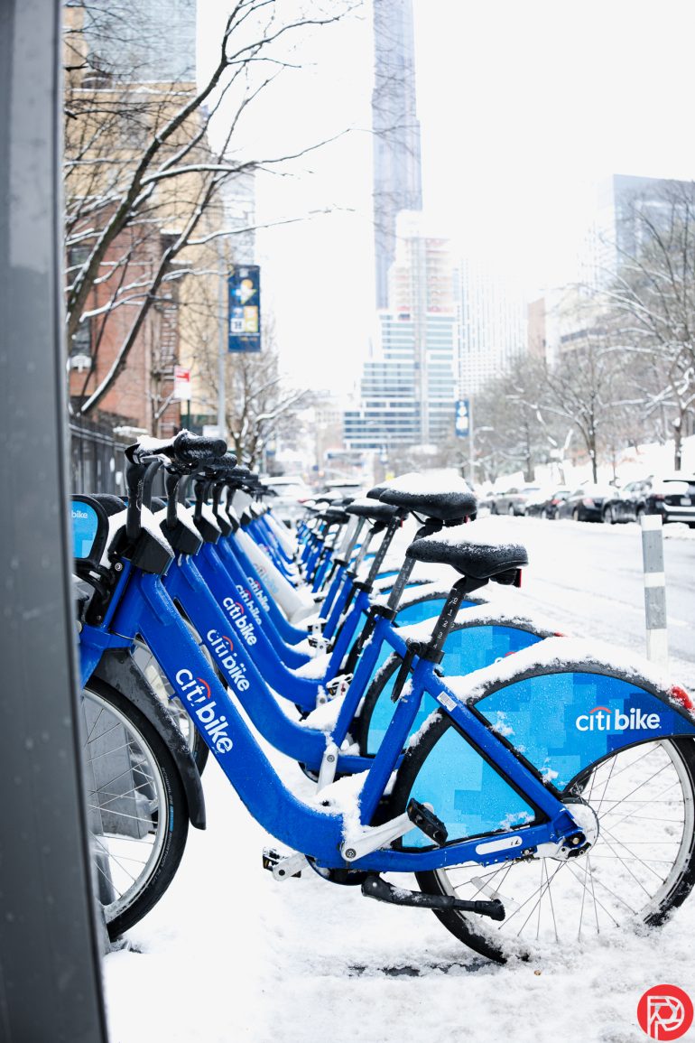 Row of blue Citi Bikes covered in snow on a snowy urban street with tall buildings in the background.