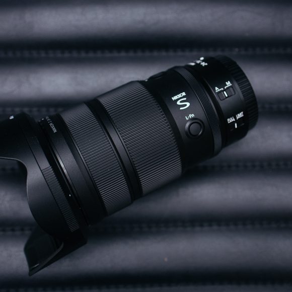 A black camera zoom lens with a hood rests on a dark, ridged surface; red logo in the corner.