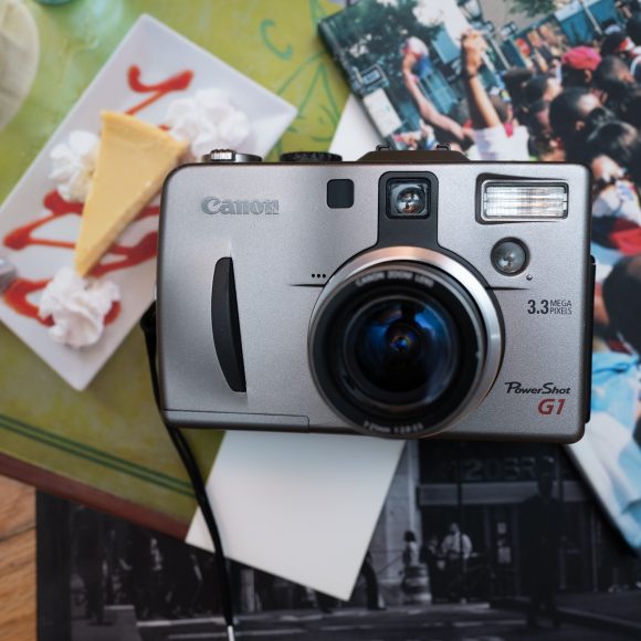 Canon PowerShot G1 camera on magazines with cheesecake and crowd photos on a wooden surface.