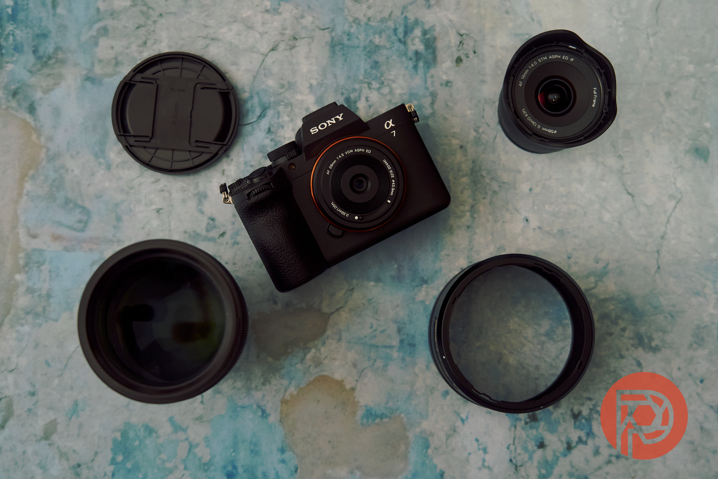 Sony camera and three lenses arranged on a blue surface, with lens caps nearby and a red logo in the corner.