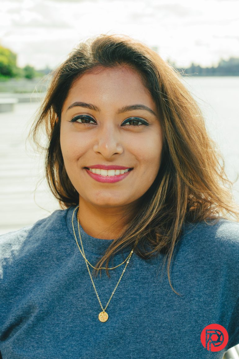 A woman with long brown hair smiles outdoors by the water, wearing a blue shirt and gold necklaces.
