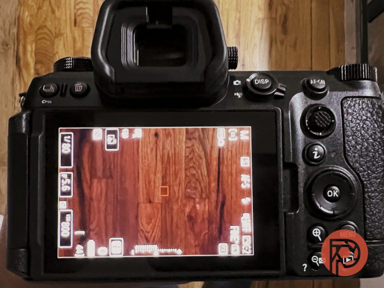 A digital camera screen displays a focus frame over a wooden floor with various camera settings visible.
