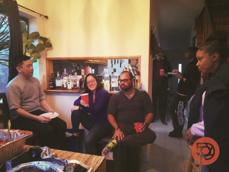 Five people with drinks gather in a kitchen, talking and relaxing near a counter with bottles.