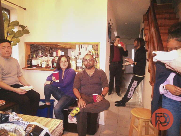 Six adults gather in a cozy kitchen, some seated with drinks, others standing and chatting by the stairs.
