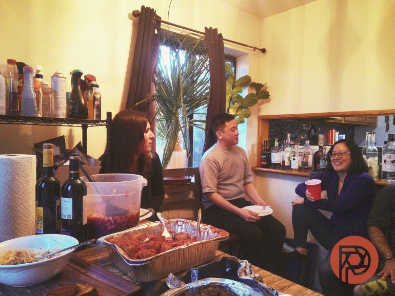 Three people sit and chat in a kitchen with food trays and drinks on the counter.