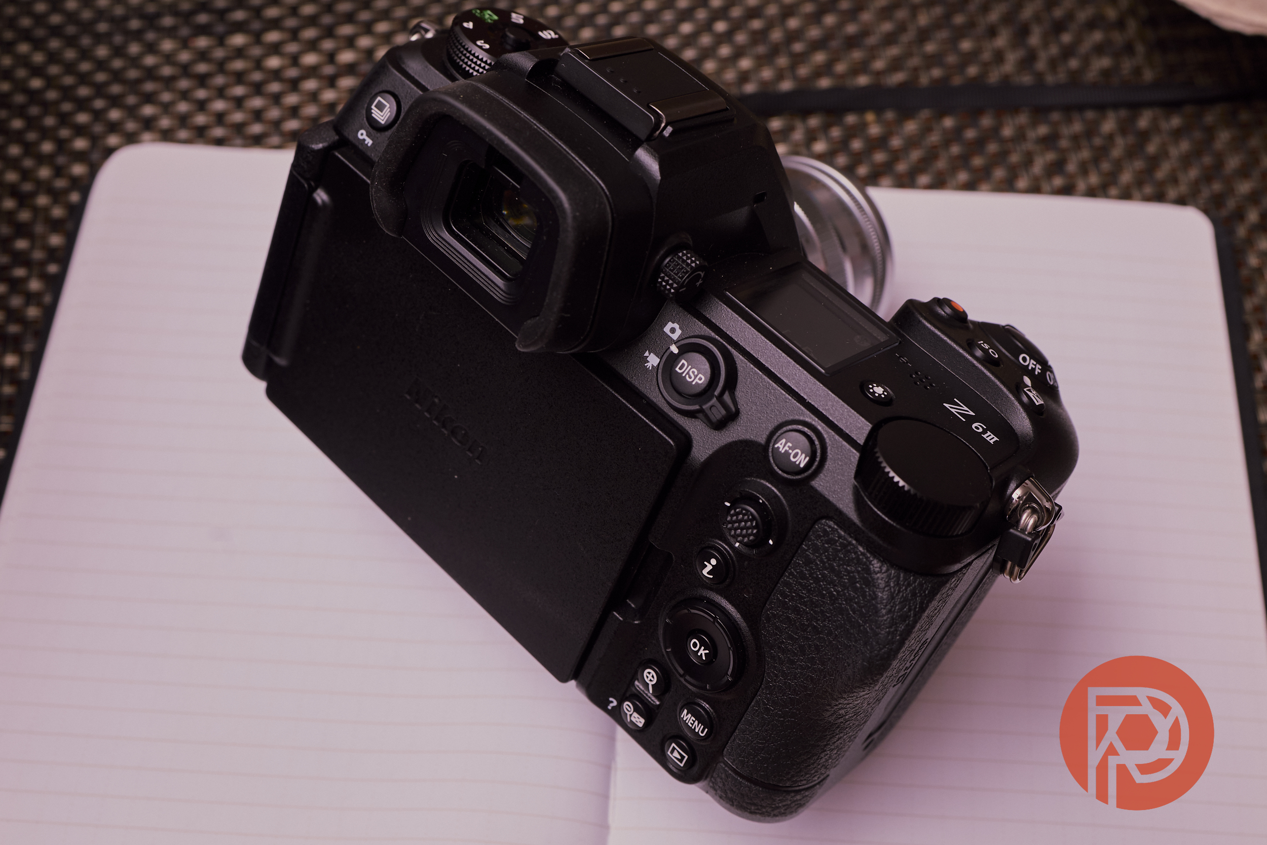 A black digital camera rests on an open blank notebook with a textured background and a red circular logo.