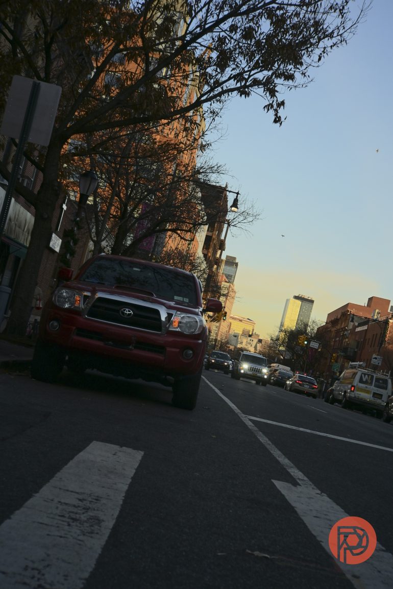 A red Toyota truck is parked on a city street at sunset, with buildings and cars in the background.