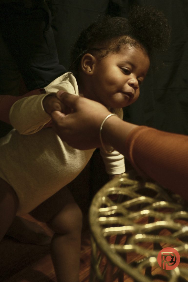 A baby in a light onesie holds an adult’s hand, leaning forward near a gold-colored basket.