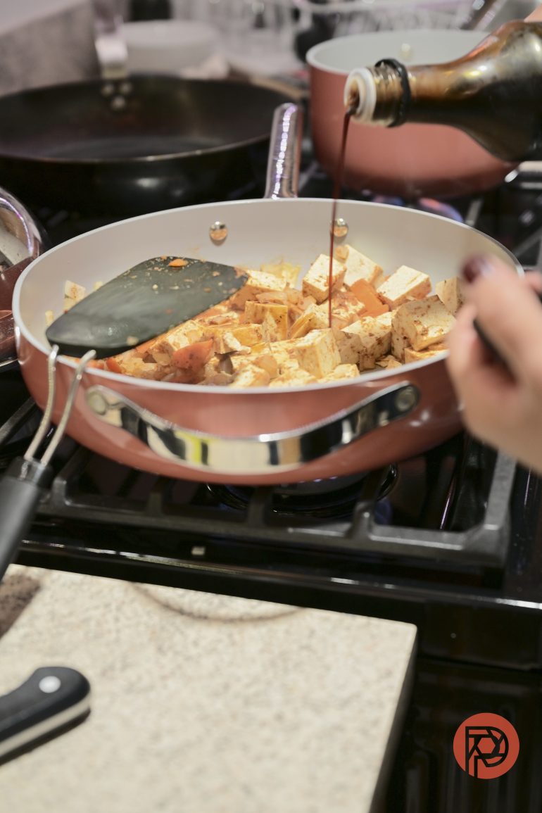 Hand pours sauce over diced tofu in a frying pan on a stove, with a spatula nearby.