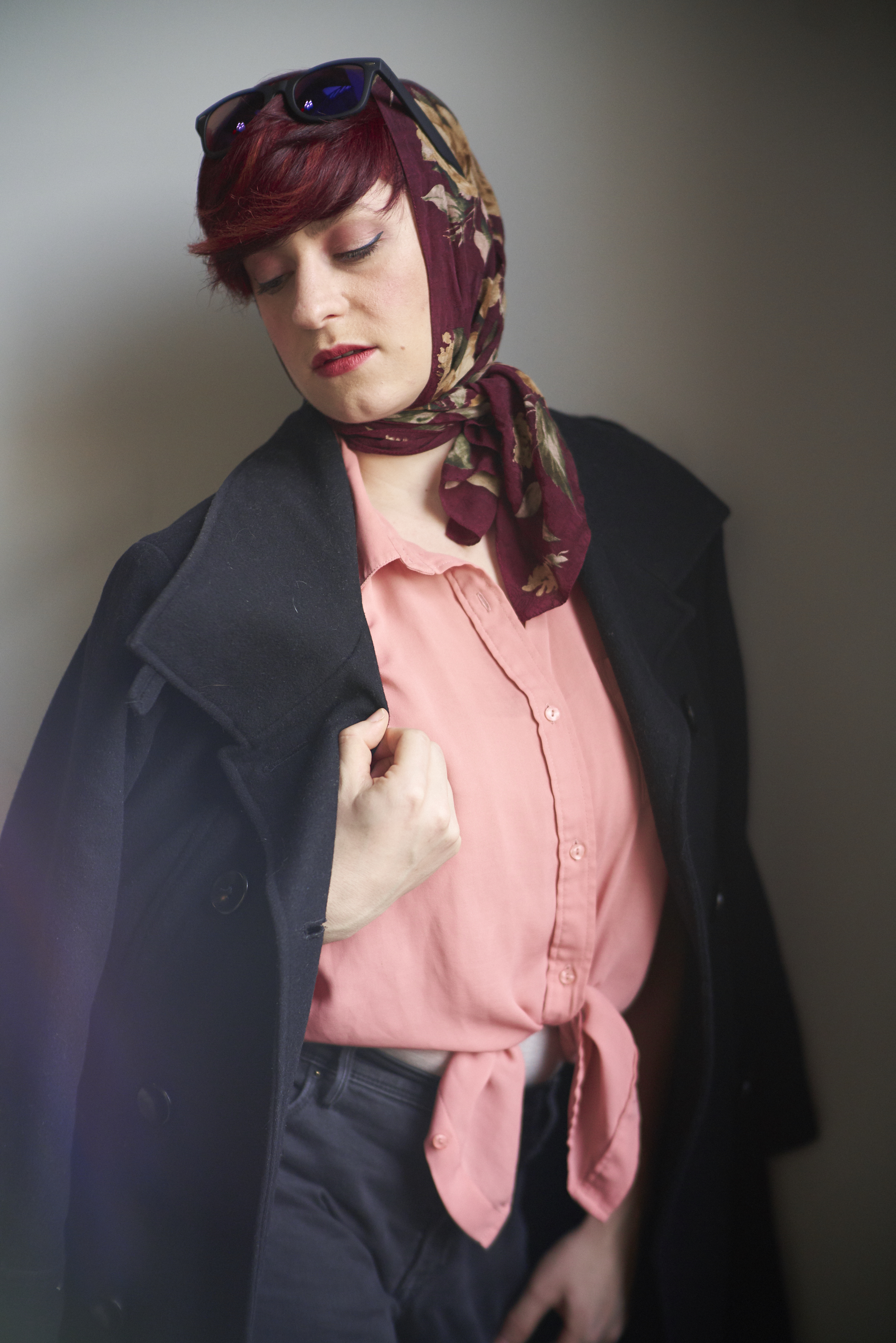 Person with short red hair, wearing a headscarf, pink shirt, and black coat, looking down.