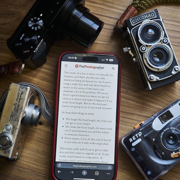 A smartphone displays an article on a desk surrounded by four vintage cameras.