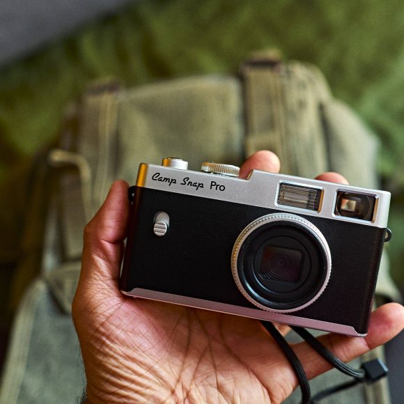 A hand holding a Camp Snap Pro camera above a green backpack.