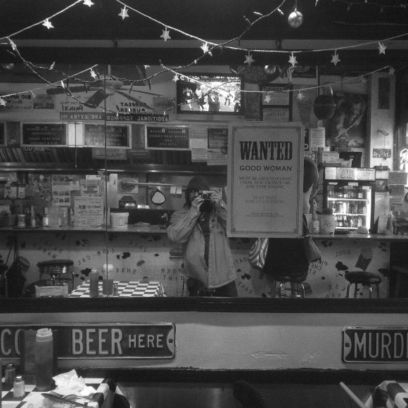 Person taking a photo in a diner with vintage signs, string lights, and a WANTED poster reflected in a mirror.
