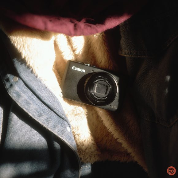 A Canon digital camera rests on a fluffy beige surface, surrounded by dark and maroon fabric in soft sunlight.
