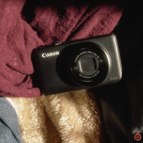 A black Canon PowerShot S95 camera resting on a maroon cloth and beige textured fabric in sunlight.