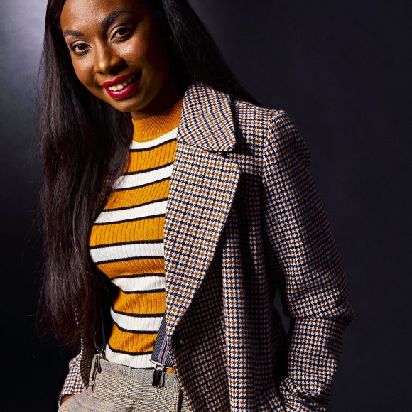 Person in a houndstooth coat and striped shirt smiles against a dark background.