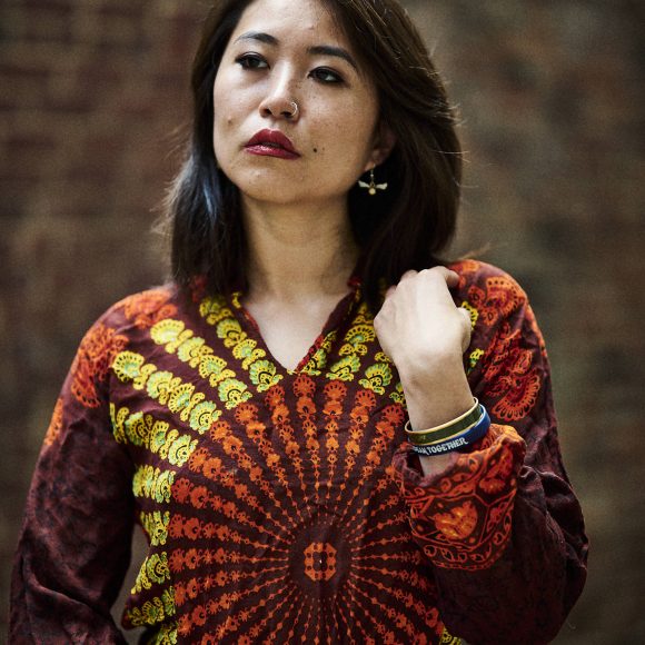 Woman in a vibrant, patterned shirt stands against a brick wall, looking upward thoughtfully.