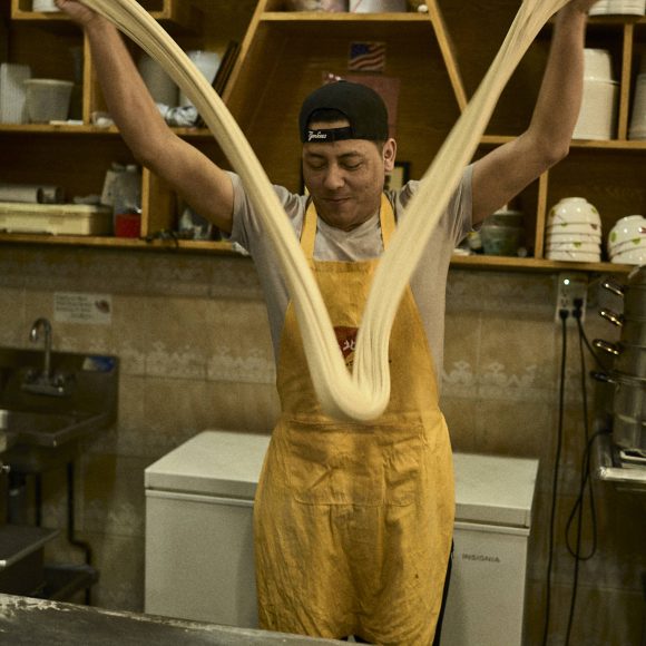 Chef stretching pizza dough in a kitchen wearing a yellow apron and cap, with shelves and clock in the background.