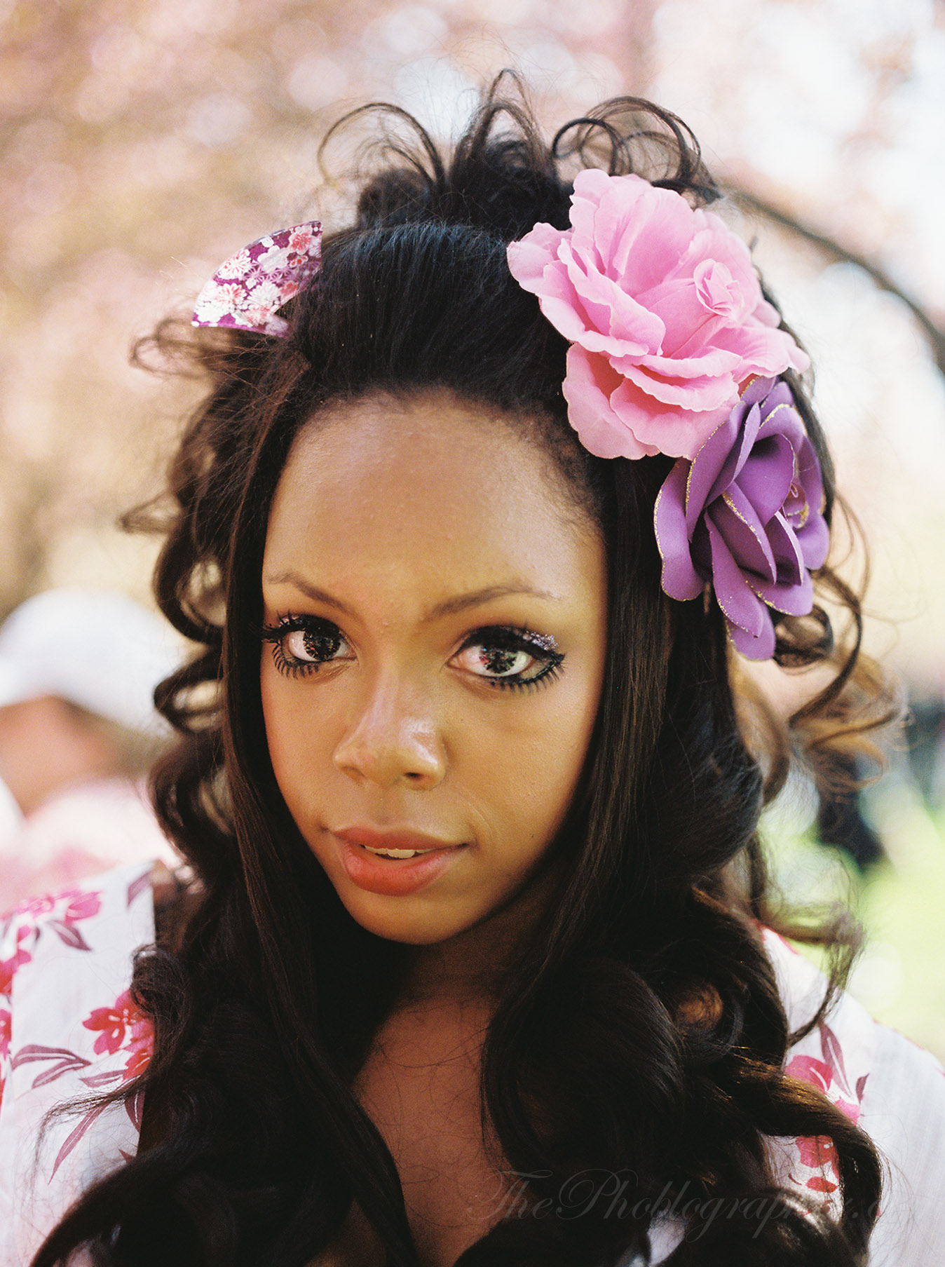 Woman with long curly hair adorned with pink and purple flowers, standing outdoors on a sunny day.