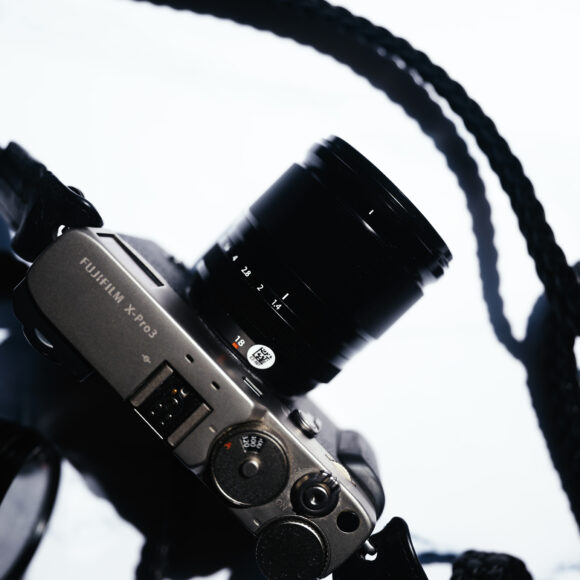 A Fujifilm X-series camera with a lens and black strap on a white surface, photographed from above.