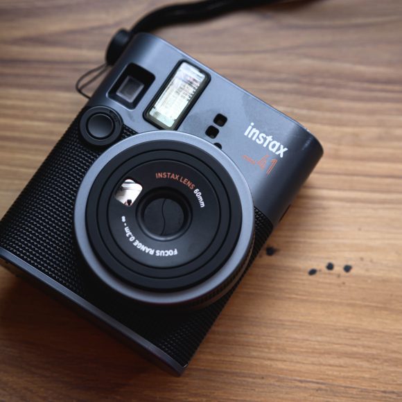 Black Instax camera placed on a wooden surface, showing its lens and flash.