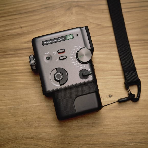 Fujifilm Instax Mini Evo Cinema Review: Why You Shouldn't Get it A black handheld electronic device with dials and buttons on a wooden surface, attached to a black strap.