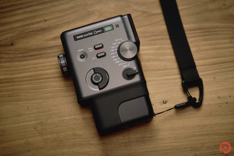 A black instant film camera with strap lies on a wooden surface, showing dials and buttons.