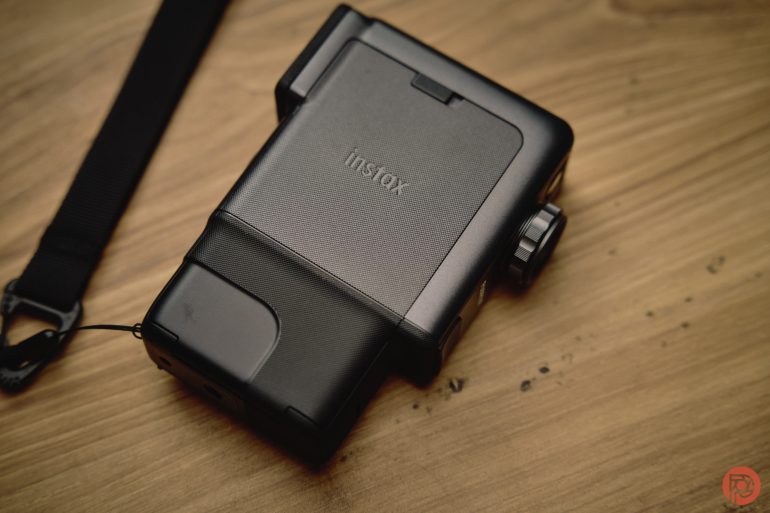 A black Instax instant camera on a wooden surface with part of its strap visible on the left.