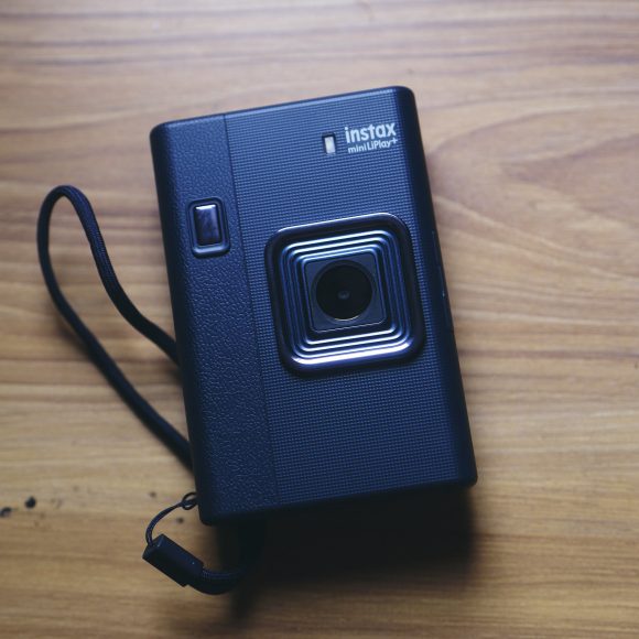 A black Instax Mini Evo camera with a wrist strap on a wooden surface.