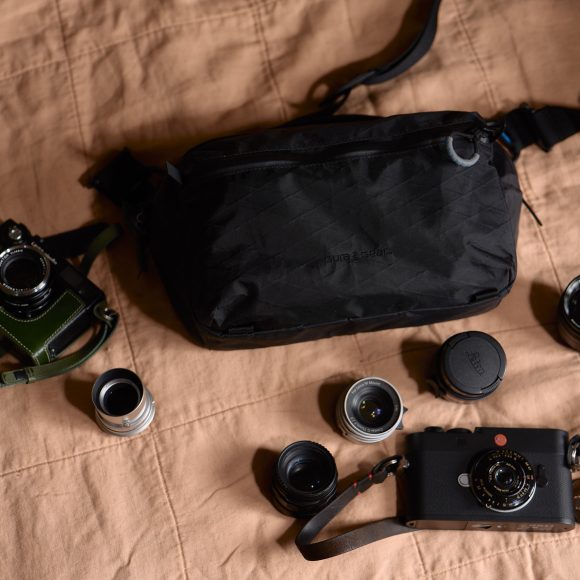 Two cameras, several camera lenses, and a black shoulder bag arranged on a peach-colored quilted surface.