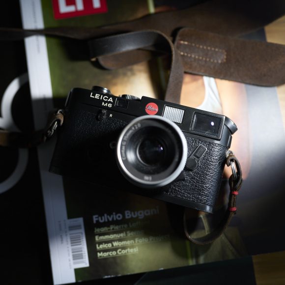A Leica M6 camera with a leather strap resting on top of magazines on a table.