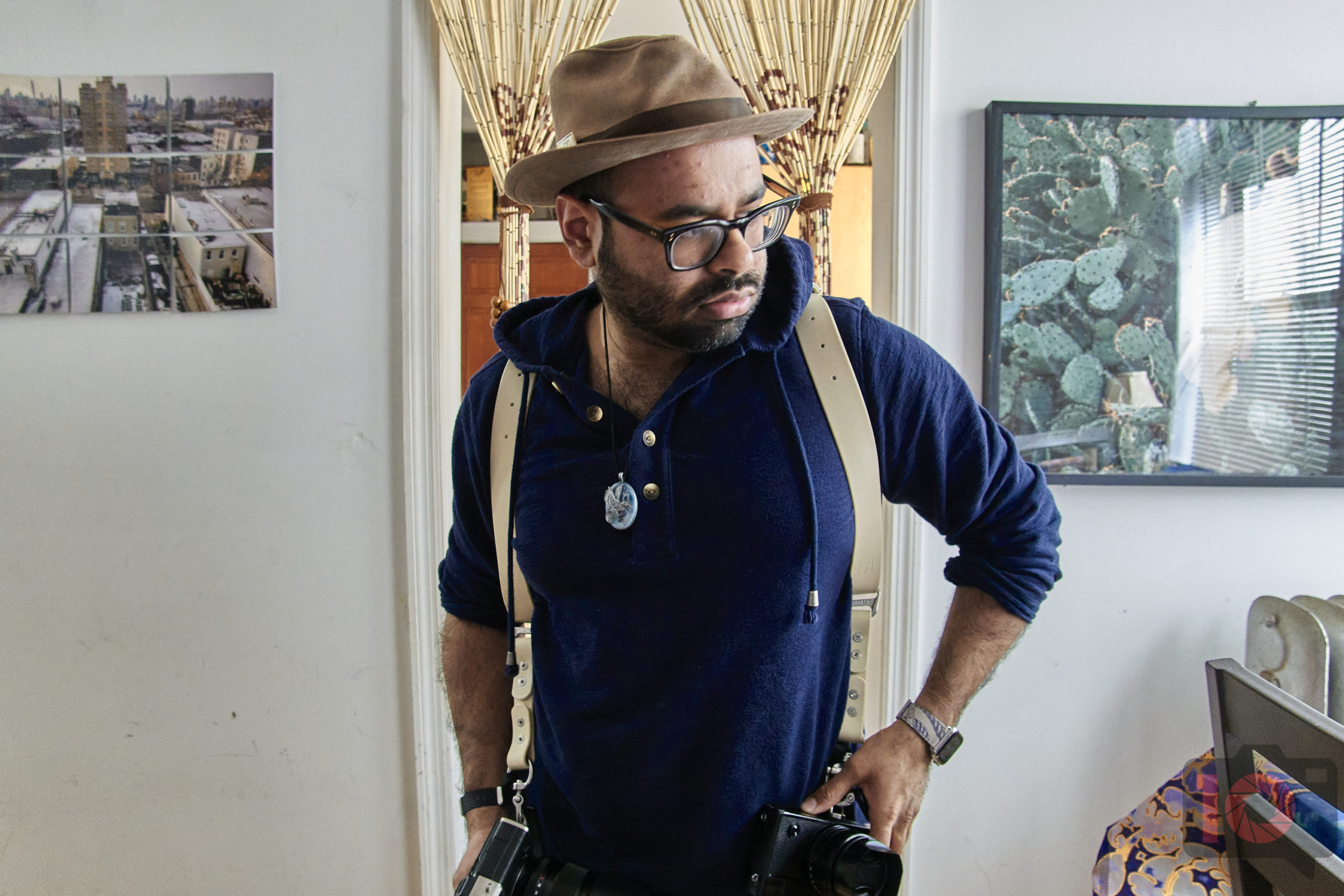 A person wearing a hat and glasses stands indoors with cameras. Wall art and a plant poster are visible in the background.