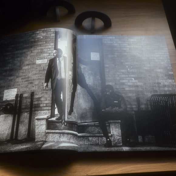 Open photobook on a wooden table showing two men near a brick building, one seated and one standing outside.
