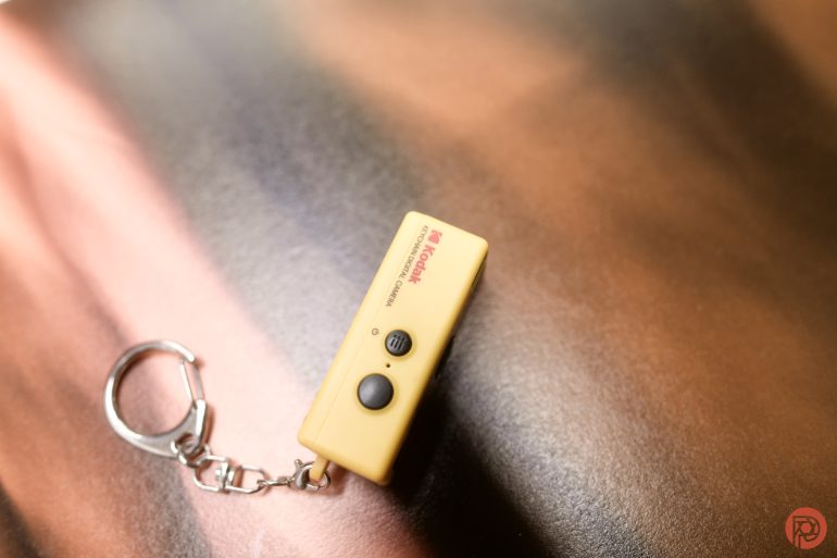 A yellow Kodak remote control keychain rests on a blurred brown and pink surface.
