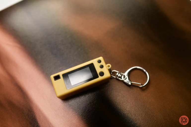 A yellow digital keychain device with a screen lies on a brown and black blurred surface.