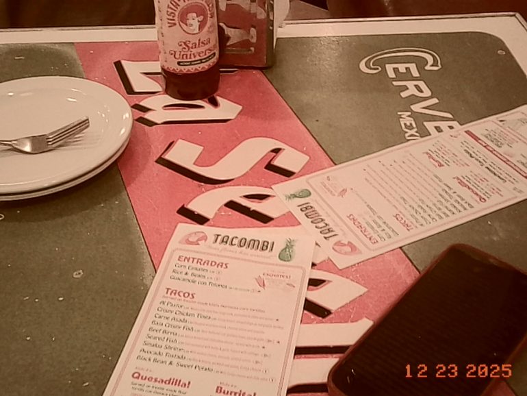 Restaurant table with menus, a salsa bottle, a phone, and a plate with utensils; date stamp: 12-23-2025.