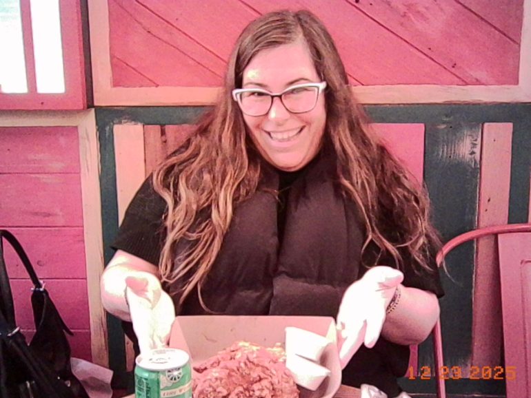 Smiling woman with glasses presenting a plate of food and a drink at a colorful restaurant table.