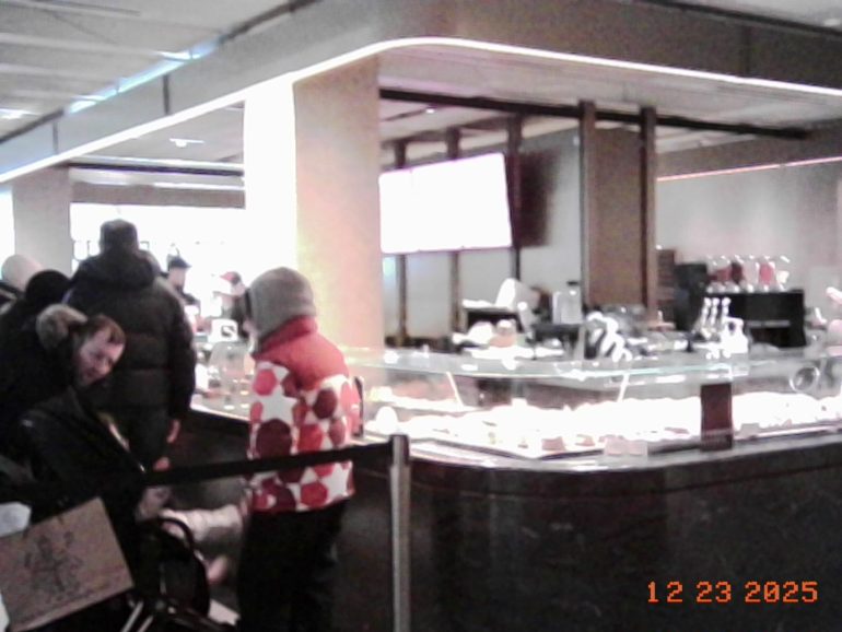 People wait in line at a food counter inside a modern, well-lit building. Date stamp: 12/23/2025.