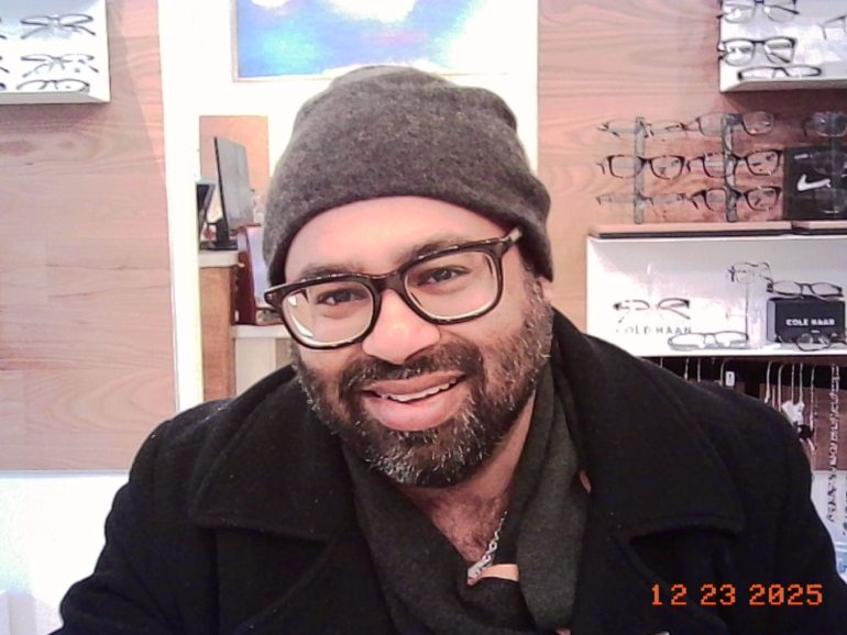 A bearded person in glasses and a beanie smiles in an eyewear store, dated 12-23-2025.