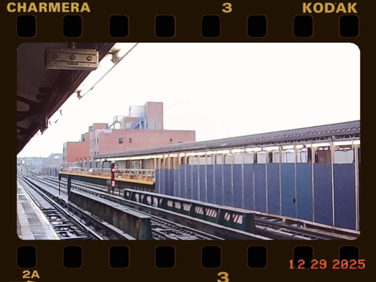 View of an elevated subway platform with tracks, buildings, and a yellow train under cloudy skies.