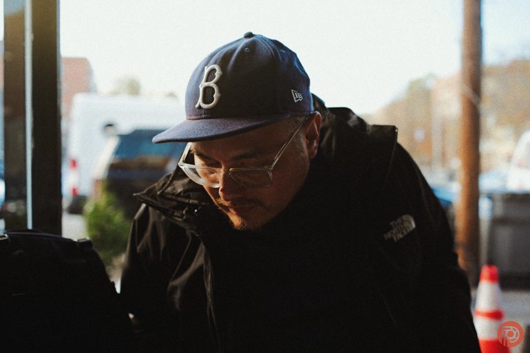 A man in a blue cap and glasses looks down, wearing a black jacket outdoors on a sunny day.