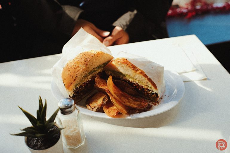 A plate with a burger cut in half and thick potato wedges on a white table, with a salt shaker and a small plant nearby.