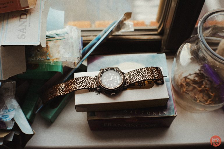 A wristwatch with a textured band rests on a box by a cluttered windowsill with scattered items.