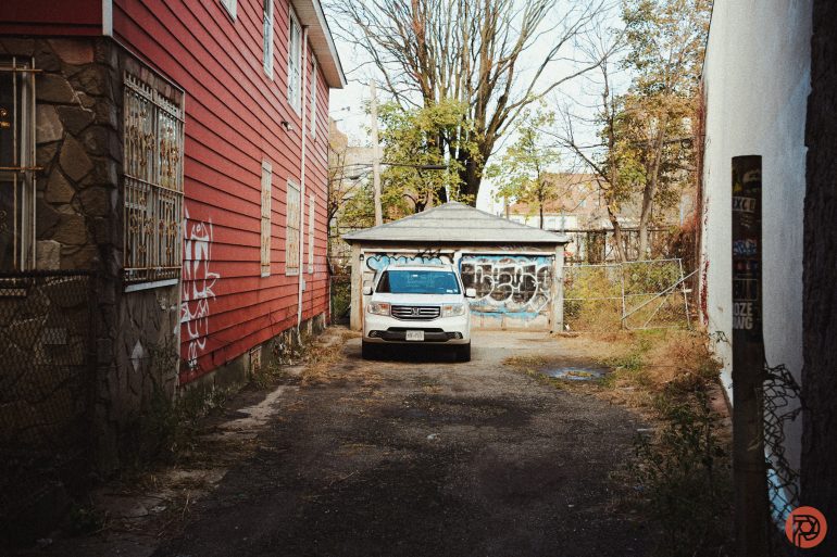 A white SUV is parked in front of a graffiti-covered garage between two buildings in an alleyway.