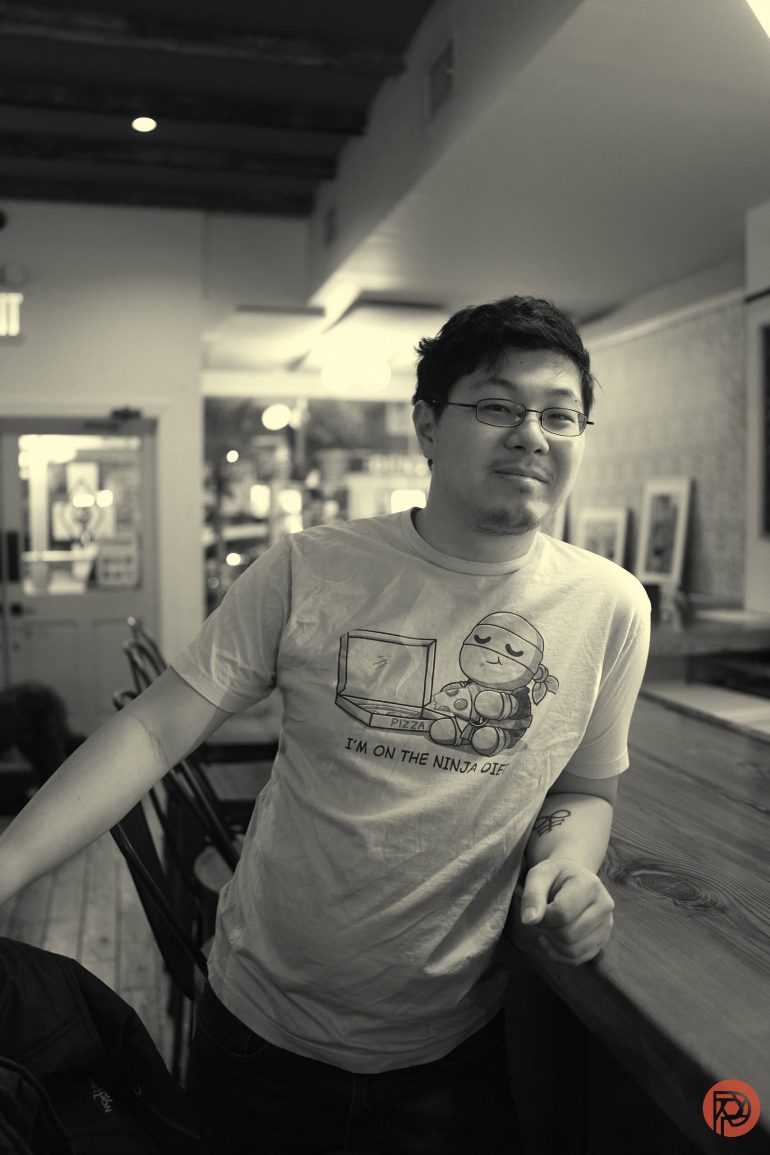 Person with glasses leans against a wooden counter in a cozy, softly lit cafe, wearing a graphic t-shirt.