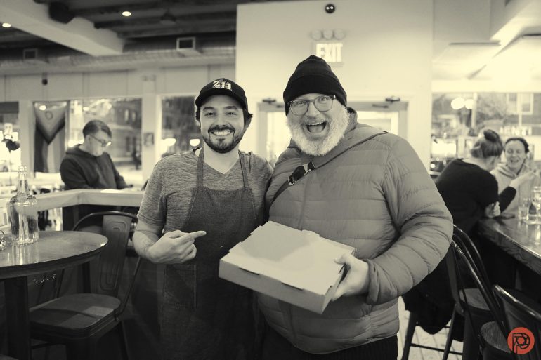 A smiling chef and a happy customer in a beanie hold a pizza box inside a busy restaurant.