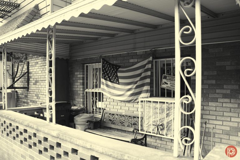 Black-and-white photo of a porch with a U.S. flag, bench, trash bins, and a For Sale sign in the window.
