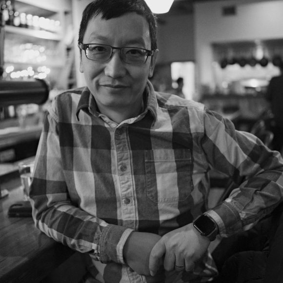 A person in glasses sits at a bar, smiling, in a black and white photo with a relaxed atmosphere.